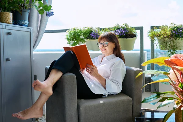 Middle-aged relaxed happy woman reading book, sitting in chair on outdoor balcony terrace at home. Rest, relaxation, calm, lifestyle, leisure, reading, mature people concept