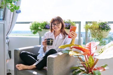 Orta yaşlı, mutlu bir kadın sandalyesinde oturmuş elinde bir fincan akıllı telefonla kameraya bakarak güneşli bir balkon terasında dinleniyor. Dinlenme, rahatlama, sakinlik, yaşam tarzı.