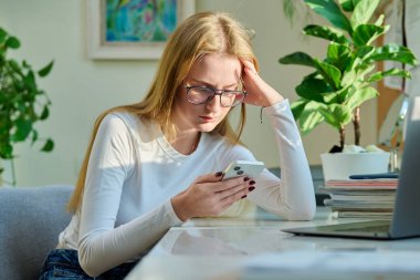 Ciddi üzgün genç bayan evde oturup akıllı telefondan mesaj okuyor. Sosyal hayat, iletişim, eğitim, yaşam tarzı teknolojisi kavramı