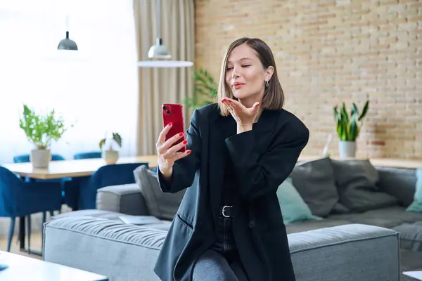 Genç, güler yüzlü, mutlu bir kadın oturma odasında kanepede oturuyor. Görüntülü konuşma görüşmesi yapıyor. Akıllı telefon kullanıyor, kameraya öpücük yolluyor. İletişim eğlence blogu vlog yaşam tarzı gençlik teknolojisi