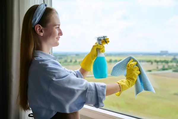 Young woman cleaning windows using spray and rag, window in an apartment home, copy space sky. Homework, housework, cleaning service, cleanliness concept