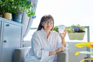 Bir bardak suyla gülümseyen orta yaşlı bir kadının portresi kameraya bakıyor, sandalyesinde güneşli teras balkonunda. Sağlık, yiyecek içecekleri, diyet, olgun insanlar konsepti