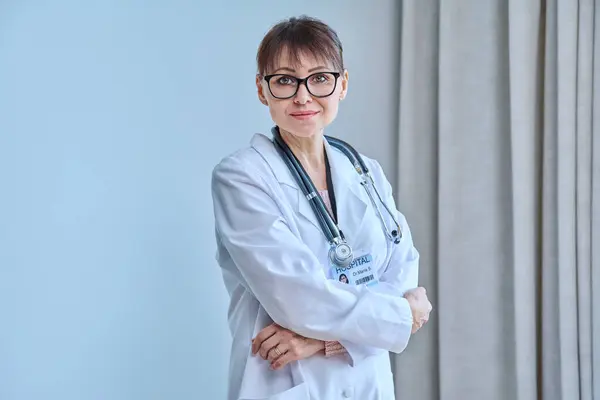 Portrait of confident mature female doctor looking at camera with arms crossed in office. Middle age woman in white coat with stethoscope with hospital medic ID card. Medicine staff service healthcare