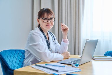 Dost canlısı, olgun bir kadın doktor dizüstü bilgisayarla masada oturuyor ve kameraya bakıyor. Beyaz önlüklü orta yaşlı kadın steteskop ve hastane kimliği. Tıbbi hizmet ilacı.