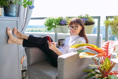 Orta yaşlı, mutlu bir kadın sandalyesinde oturmuş elinde bir fincan akıllı telefonla kameraya bakarak güneşli bir balkon terasında dinleniyor. Dinlenme, rahatlama, sakinlik, yaşam tarzı.