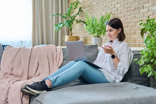 Young relaxed woman sitting on sofa at home in living room, using laptop for video call conference chat. Holidays, leisure, technology, comfort, lifestyle, youth concept