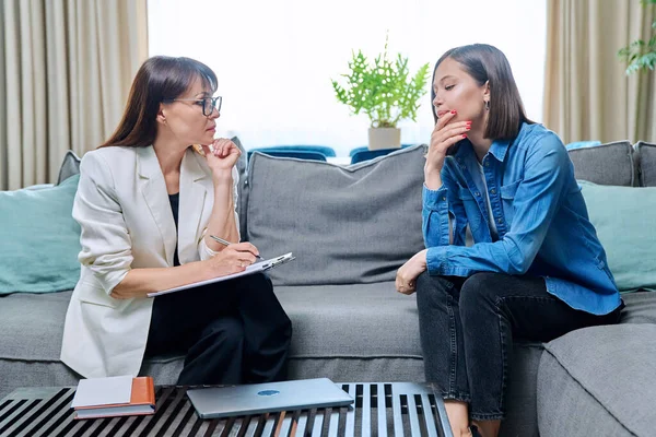 Sad young 20s woman in meeting with female psychologist therapist counselor sitting on couch in office. Psychology, therapy, psychotherapy, treatment, mental health concept