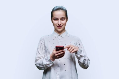 Genç bir öğrenci, 15-16 yaşlarında, akıllı telefonlu bir kız beyaz stüdyo arka planında kameraya bakıyor. Ergenlik, internet teknolojileri, mobil çevrimiçi uygulamalar boş zamanları öğrenmek için uygulama hizmetleri