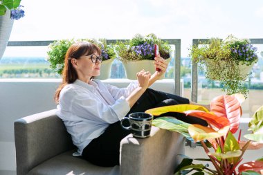 Orta yaşlı, gevşemiş, sohbet konferansı için akıllı telefon kullanan, balkon terasında oturan olgun bir kadın. Dinlenme rahatlama yaşam tarzı rahatlama iletişimi