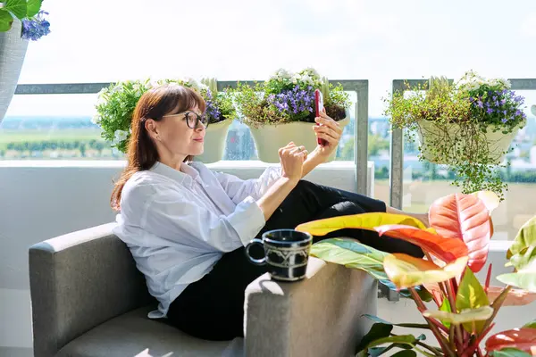 Orta yaşlı, gevşemiş, sohbet konferansı için akıllı telefon kullanan, balkon terasında oturan olgun bir kadın. Dinlenme rahatlama yaşam tarzı rahatlama iletişimi