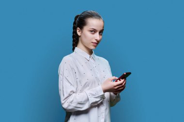 Genç bir öğrenci, 15-16 yaşlarında, akıllı telefonlu bir kız mavi stüdyo arka planındaki kameraya bakıyor. Ergenlik, internet teknolojileri, mobil çevrimiçi uygulamalar boş zamanları öğrenmek için uygulama hizmetleri