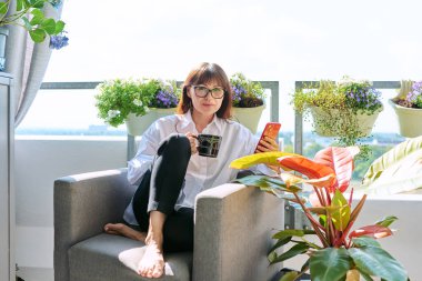 Orta yaşlı, mutlu bir kadın sandalyesinde oturmuş elinde bir fincan akıllı telefonla kameraya bakarak güneşli bir balkon terasında dinleniyor. Dinlenme, rahatlama, sakinlik, yaşam tarzı.