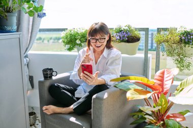 Orta yaşlı, gevşemiş, sohbet konferansı için akıllı telefon kullanan, balkon terasında oturan olgun bir kadın. Dinlenme rahatlama yaşam tarzı rahatlama iletişimi