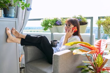Orta yaşlı, mutlu bir kadın sandalyesinde oturmuş cep telefonuyla konuşuyor, güneşli bir balkon terasında dinleniyor. Dinlenme, rahatlama, yaşam tarzı, rahatlık, iletişim, olgun insanlar.