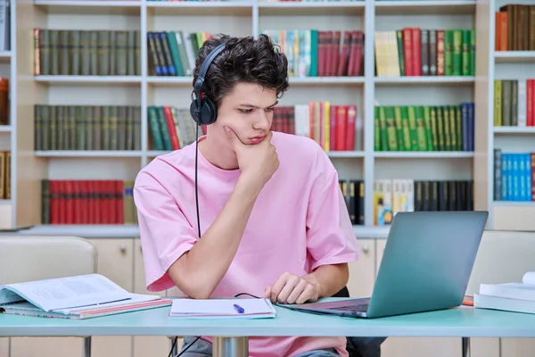 Young male student sitting with computer laptop books in college library. Guy 18-20 years old with headphones typing on laptop. Knowledge, education, youth, college university concept