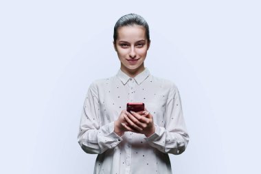 Genç bir öğrenci, 15-16 yaşlarında, akıllı telefonlu bir kız beyaz stüdyo arka planında kameraya bakıyor. Ergenlik, internet teknolojileri, mobil çevrimiçi uygulamalar boş zamanları öğrenmek için uygulama hizmetleri