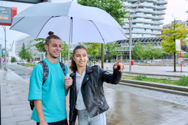 Otobüs durağında şemsiyenin altında sırt çantalı genç bir erkek ve kız öğrenci belediye otobüsünü bekliyor. Toplu taşıma, ergenlik, gençlik, şehir hayatı, yaşam tarzı, şehir tarzı.
