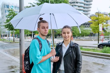 Modern şehrin sokaklarında şemsiye altında kameraya bakan genç öğrencilerin, gülümseyen erkek ve kızların açık hava portresi. Üniversite, lise, şehir yaşam tarzı, ergenlik, gençlik konsepti