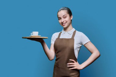 Mavi stüdyo arka planında bir tepsi kahveyle önlüklü genç bayan garson portresi. Gülümseyen kız kameraya bakıyor. İş, yemek servisi, gençlik kavramı