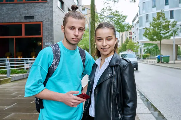 Outdoor portrait of teenagers students friends smiling guy and girl with smartphones looking at camera on street of modern city. College, 17-19 years old, urban lifestyle, adolescence youth concept