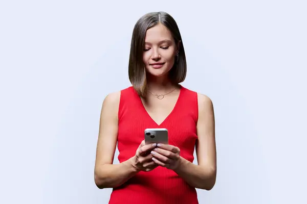 Young woman with smartphone in hands on white studio background. Smiling female looking at phone screen texting reading. Using mobile applications for work business shopping banking study leisure