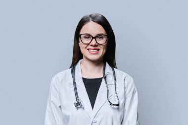 Beyaz önlüklü gülümseyen kadın doktorun portresi gri arka planda kameraya bakıyor. Tıbbi personel, meslek, sağlık hizmetleri, bilim, tıp konsepti