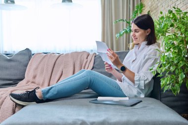 Gülümseyen mutlu genç kadın resmi kağıt mektup okuyor, evdeki oturma odasında kanepede oturuyor. Banka, iş, sosyal vergi hizmetleri, fatura, sigorta şirketi, kiralık mallar hakkında bilgilendirme
