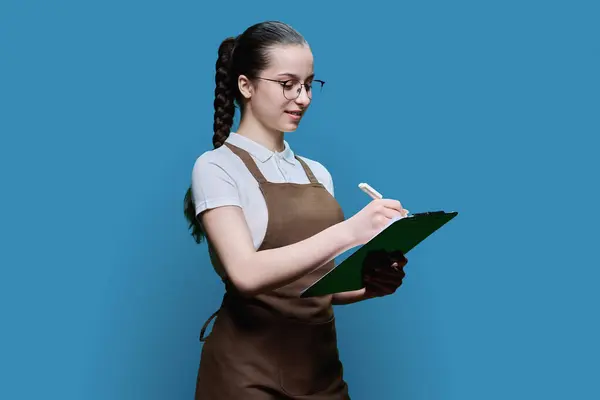 Young female worker in apron writing on clipboard paper, on blue background. Work, service, small business, youth concept