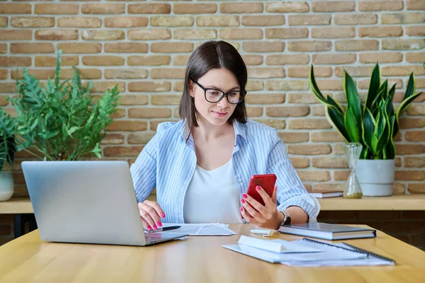 stock image Young business woman sitting at desk with laptop computer in office, using smartphone. 30 years old confident female at work