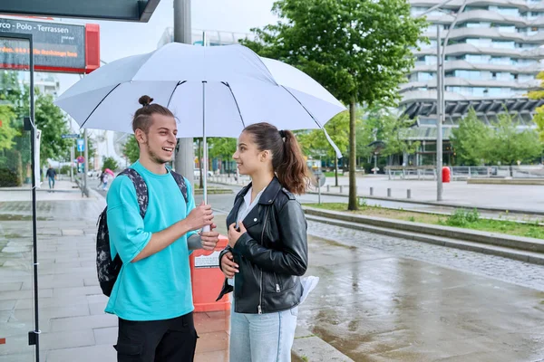 Otobüs durağında şemsiyenin altında sırt çantalı genç erkek ve kız öğrenciler yağmurlu havalarda şehir otobüsü, yol fotokopi alanı bekliyorlar. Toplu taşıma, gençlik, şehir yaşam tarzı, şehir tarzı.