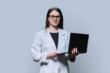 Genç kadın uzmanın portresi, profesyonel tıp bilimcisi, gri arka planda dizüstü bilgisayar kullanan beyaz önlüklü eczacı. Sağlık personeli, meslek, sağlık hizmetleri, bilim ilaçları, çevrimiçi hizmetler