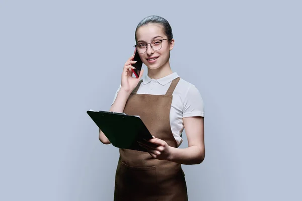 Young female worker in apron taking order by phone, writing on clipboard paper, on gray studio background. Work, service, small business, youth concept