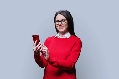 Akıllı telefonlu, kırmızı kazaklı genç bir kadın gri arka planda kameraya bakıyor. İletişim, yaşam tarzı, teknoloji mobil uygulamaları İnternet hizmetleri, ağ, 30 yaşındaki insanlar