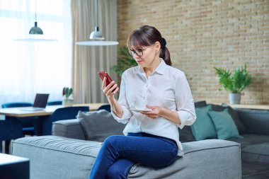 Orta yaşlı kadın akıllı telefon kullanıyor, evinde kanepede kahve içiyor. Olgun kadın dinlenme, okuma haberleri, sosyal ağlar, arkadaşlarla sohbet, iş arkadaşları, aile, mobil uygulamalar, boş zaman
