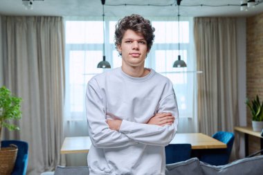 18-19 yaşlarında ev içinde çapraz kollu yakışıklı bir adamın portresi. Beyaz elbiseli kıvırcık, kendine güvenen bir üniversite öğrencisi kameraya bakıyor. Gençlik, yaş, yaşam tarzı konsepti