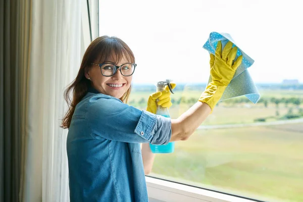 戴着手套的中年妇女 家里有喷雾式的清洁窗户 微笑着的女性看着相机 家庭清洁 打扫房屋 家庭生活 生活方式概念 — 图库照片