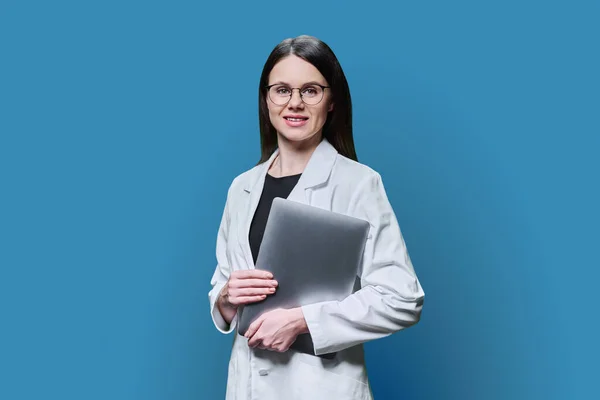 Portrait of young woman specialist, professional medical scientist, pharmacist in white coat with laptop on blue background. Medical staff, occupation, health care, science medicine, online services