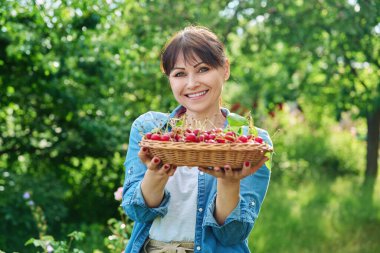 Kırmızı kiraz hasadıyla mutlu kadın yaz güneşli bahçesinde kameraya bakıyor. Hasat, tarım, bahçıvanlık, çiftçi pazarı, sağlıklı doğal vitamin organik eko meyveler, gıda kavramı.