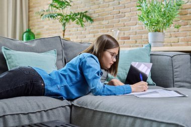 Evde ders çalışan genç bir üniversite öğrencisi dizüstü bilgisayar kullanarak kanepede uzanıyor. Öğrenim için eğitim bilgisi gençlik mobil uygulama İnternet çevrimiçi hizmet