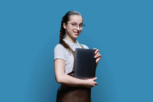 Portrait of young smiling teenage female in an apron holding laptop, looking at camera on blue studio background. Worker, small business, job, service sector, staff, youth concept
