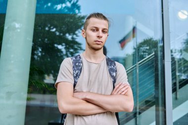 Kendine güvenen üniversite öğrencisinin portresi, kolları sırt çantasıyla bağlı, kameraya bakıyor, dış eğitim binası geçmişi var. Gençlik, eğitim, yaşam tarzı konsepti