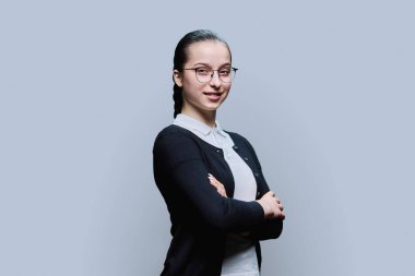 Gri arka planda gözlüklü, kendine güvenen gülümseyen genç kız portresi. Pozitif kadın, 16-17 yaşlarında çapraz kollu lise öğrencisi bir gençle kameraya bakıyor.