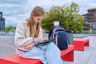 Okul bahçesinde dijital tablet kullanarak sırt çantasıyla bankta oturan ciddi bir öğrenci. Okula, çocukluğa, eğitim bilgi öğrenme kavramına