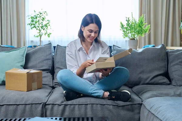 Young woman unpacking online purchase box with new smartphone while sitting on sofa in living room. Online internet shopping, postal delivery, delivery service