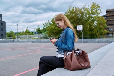 Sırt çantalı, açık havada akıllı telefon kullanan kendinden emin liseli bir kızın portresi. Gülümseyen liseli kız telefona bakıyor, okul binası geçmişi var. Ergenlik eğitimi bilgisi