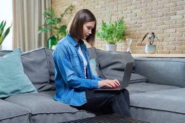 Kanepede oturan ve dizüstü bilgisayarda yazan genç bir kadın. Çalışmak için internet teknolojileri, uzaktan çalışan serbest kadın, online ders izleyen üniversite öğrencisi, e-öğrenme, dinlenme iletişimi