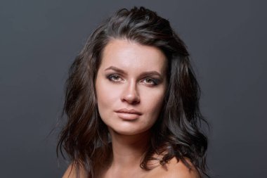 Koyu gri stüdyo arka planında, çıplak omuzlu, saç stili makyajlı genç bir kadının portresi. Kameraya bakan güzel bir esmer. Güzellik, moda, makyaj, ten, saç