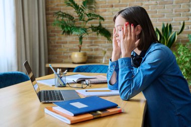 Masasında dizüstü bilgisayarı ve ders kitaplarıyla oturan üzgün, yorgun bir üniversite öğrencisi. Gençlik, eğitim, duygular depresyon yorgunluk baş ağrısı, yaşam tarzı konsepti