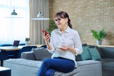 Orta yaşlı kadın akıllı telefon kullanıyor, evinde kanepede kahve içiyor. Olgun kadın dinlenme haberleri, sosyal ağlar, arkadaşlarla sohbet, iş arkadaşları, aile, sürpriz mutluluk duyguları.