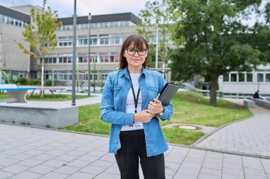 Orta yaşlı kendine güvenen kadın öğretmen, akıl hocası, pedagog, psikolog, danışman, elinde dijital tabletle okul binası yakınlarında poz veren sosyal hizmet görevlisi. Eğitim, öğretim, okul